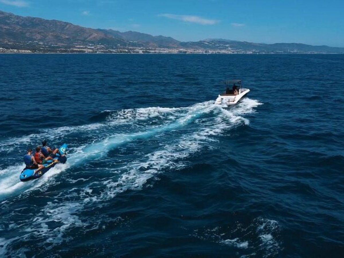 Boat towing a banana boat with people on a blue sea near a mountainous coastline.