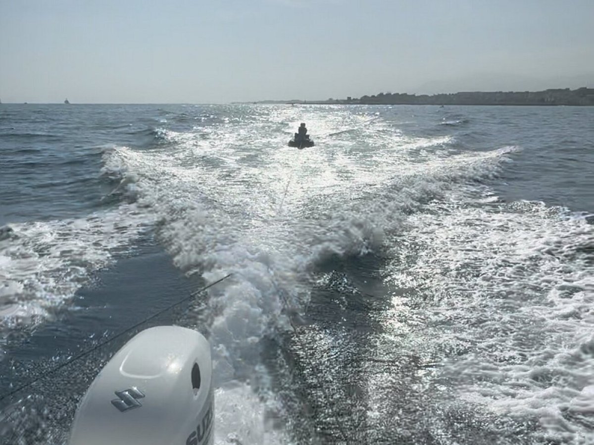 View from boat, water splashes behind, distant jet ski on ocean.