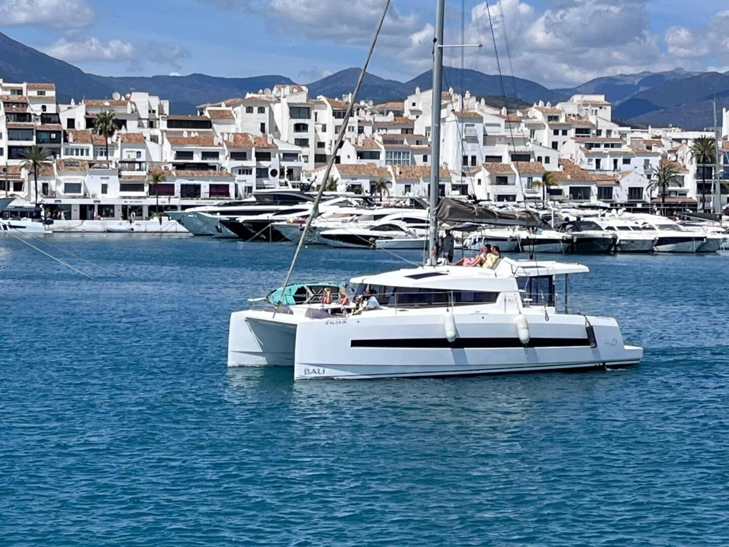 Catamaran sailing near marina with white buildings and mountains in the background.