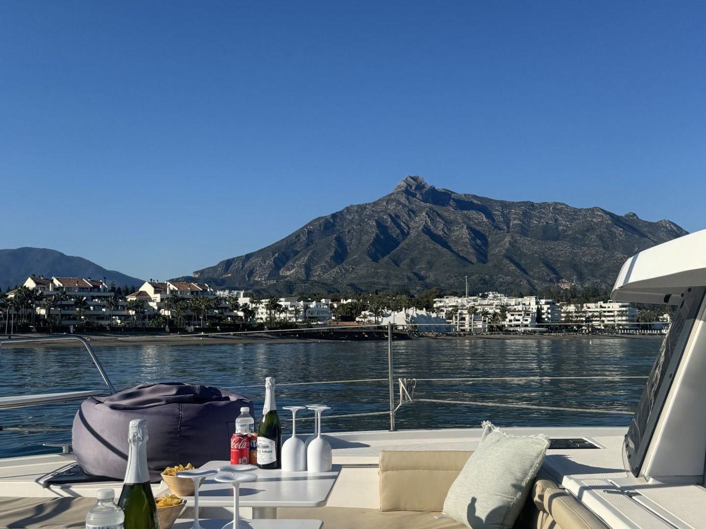 Boat deck with table, drinks, and mountain view under clear blue sky.
